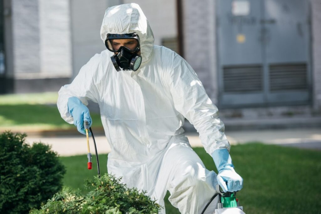 A person in protective coveralls, gloves, and a respirator mask sprays chemicals onto bushes in a garden, likely conducting pest control or disinfection.