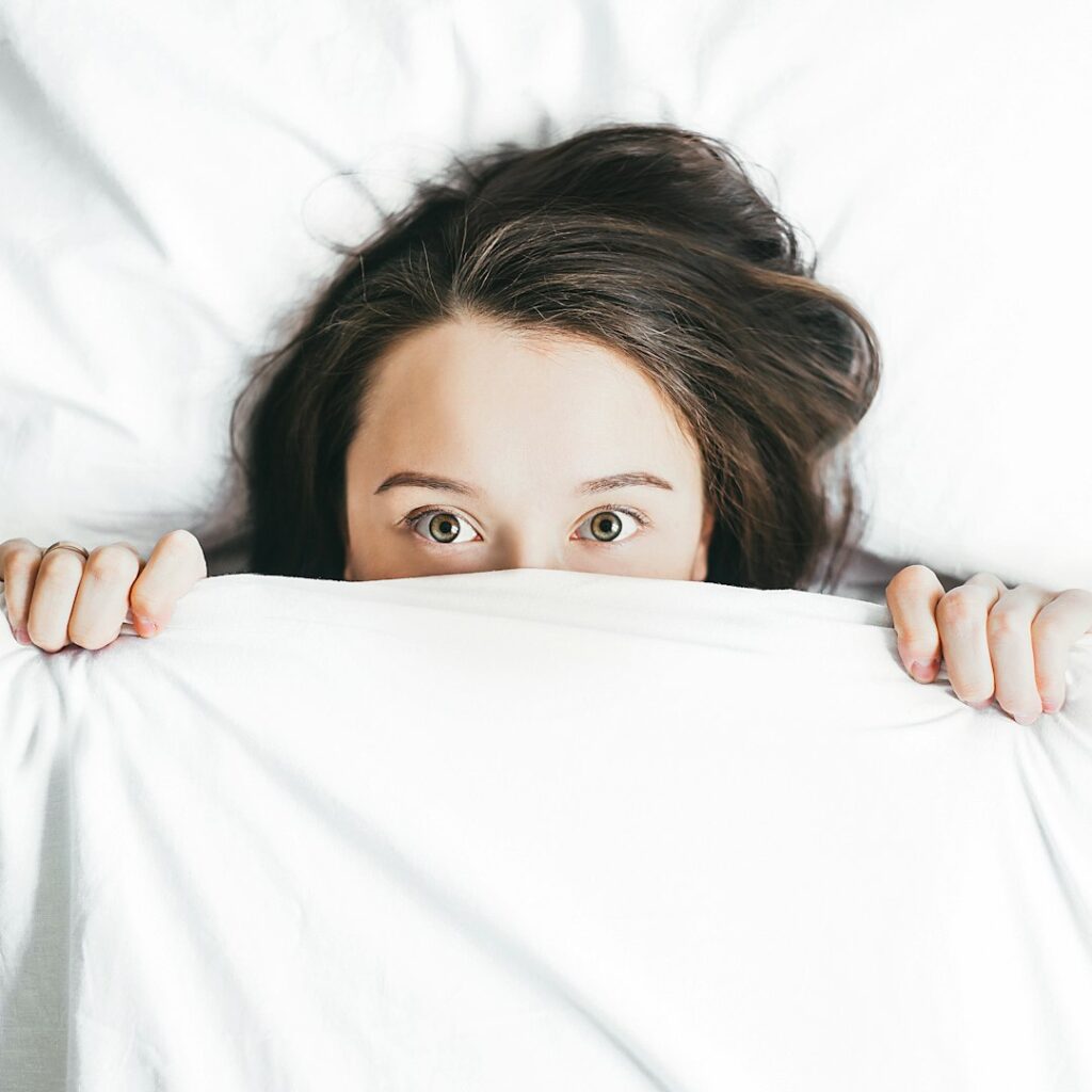 A person with long dark hair lies in bed, partially covered by a white blanket, with only their eyes and hands visible above the covers, looking directly at the camera.